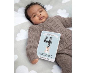 Curly-haired baby in brown sweater on blanket with cloud patterns, holding a milestone baby photo card. Photo courtesy of Milestone.